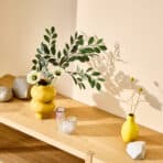 Stylish yellow vases with green foliage and decorative stones on a wooden shelf, perfect for modern home decor.