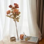 Artificial pink roses in a tall glass vase with decorative accessories by a bright window.