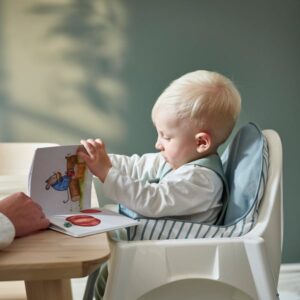 Baby in high chair reading a colorful picture book with adult hand nearby.