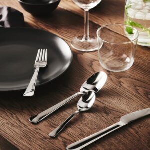 Stainless steel spoons on a wooden table with glassware and black plate.